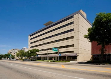 A beige multi-story office building with large windows stands beside a quiet, sunlit street in Lincoln, Nebraska, lined with trees and signs under a clear blue sky—an inviting setting near Crossroads House for independent living.