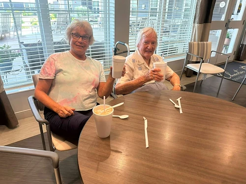 Two residents at Charter Senior Living of Hopkinsville in Hopkinsville, KY, smiling and enjoying root beer floats in the bright dining room with natural light from large windows, highlighting a joyful assisted living and memory care environment.
