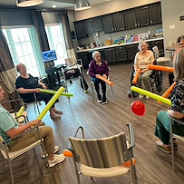 Seniors at Charter Senior Living of Hopkinsville in Hopkinsville, KY, sitting in a circle engaging in an active group activity with pool noodles and a balloon, promoting fun, fitness, and community in assisted living and memory care.